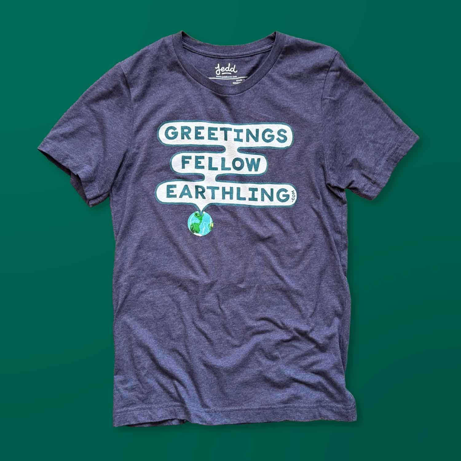 A heather blue t-shirt with a design that says Greetings Fellow Earthling