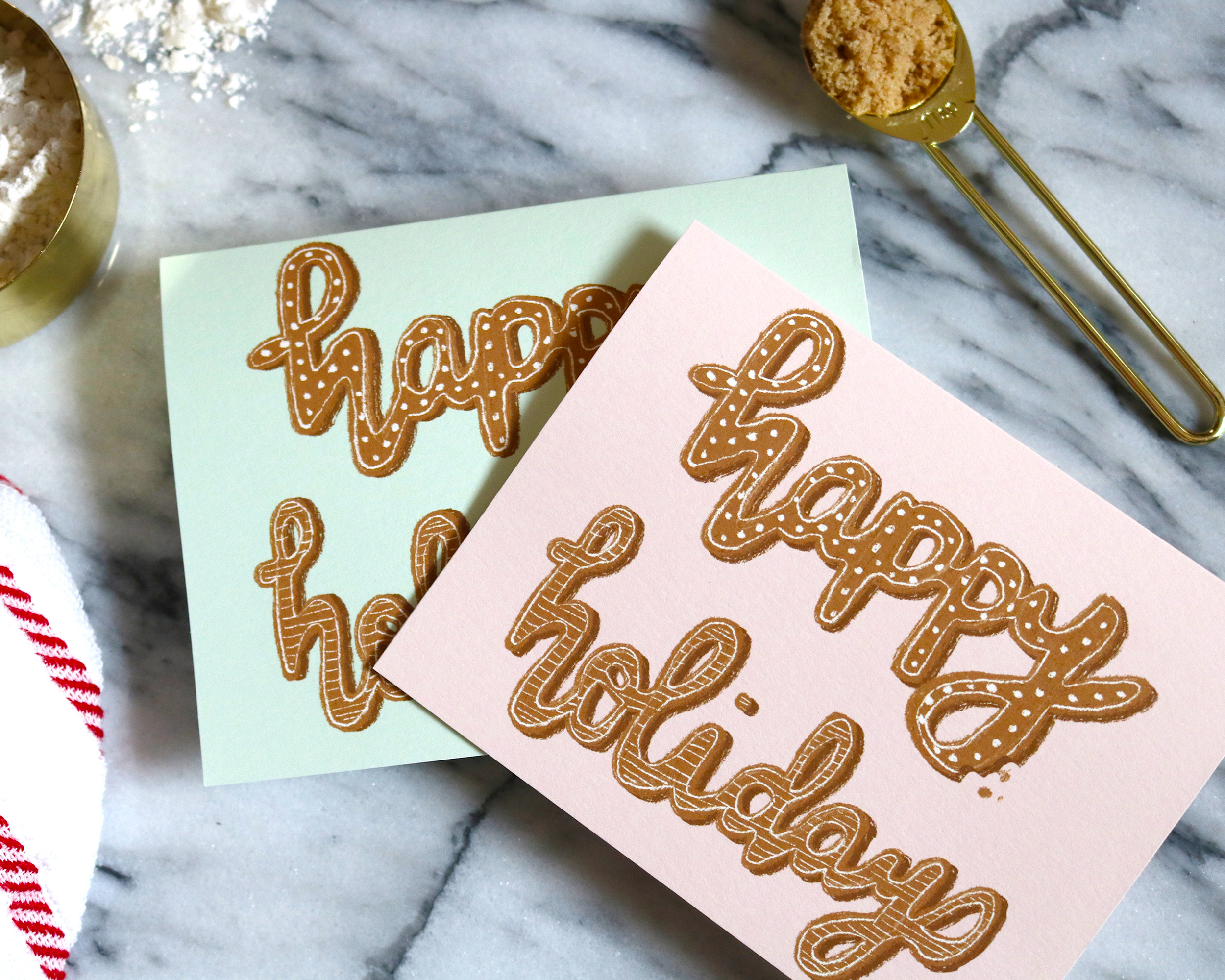 Greeting Card with illustrated happy holidays that look like frosted gingerbread cookies. Available in pink or mint. Displayed with baking supplies.