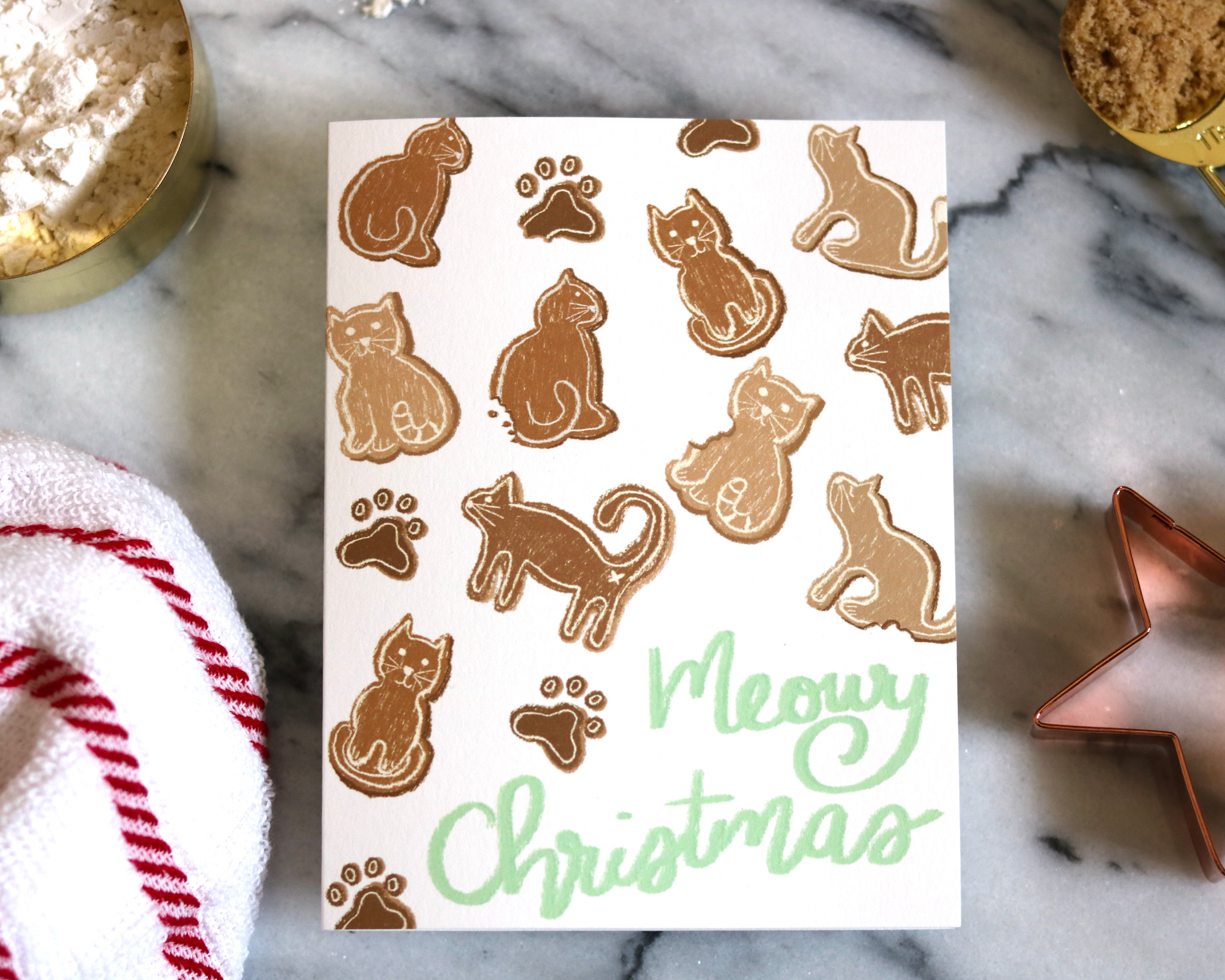 Our Meowy Christmas Gingerbread Cat Greeting card is displayed with various baking supplies on a marble background.