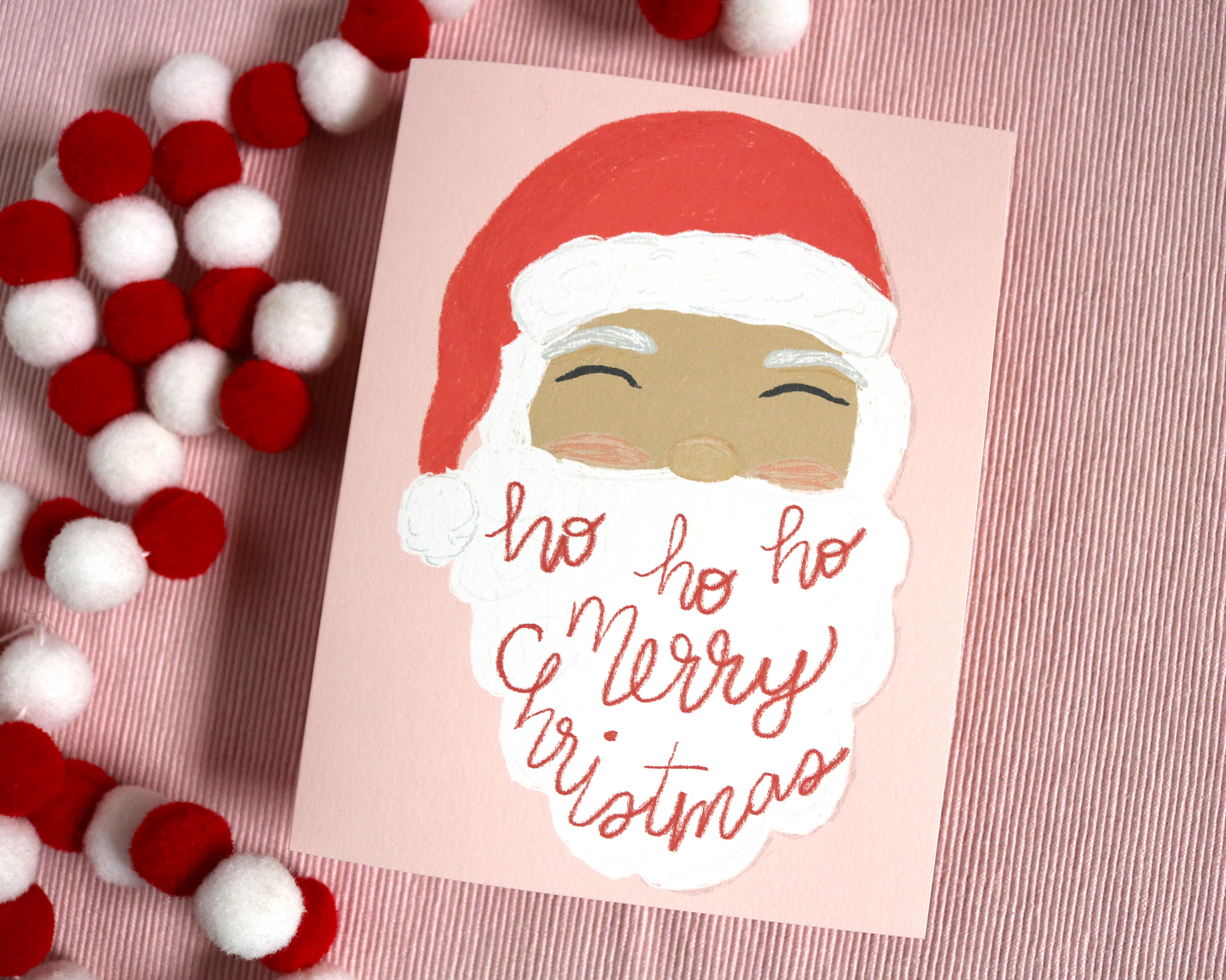 Our Vintage Inspired Ho Ho Ho Santa greeting card is displayed on pink fabric next to red and white felt ball garland