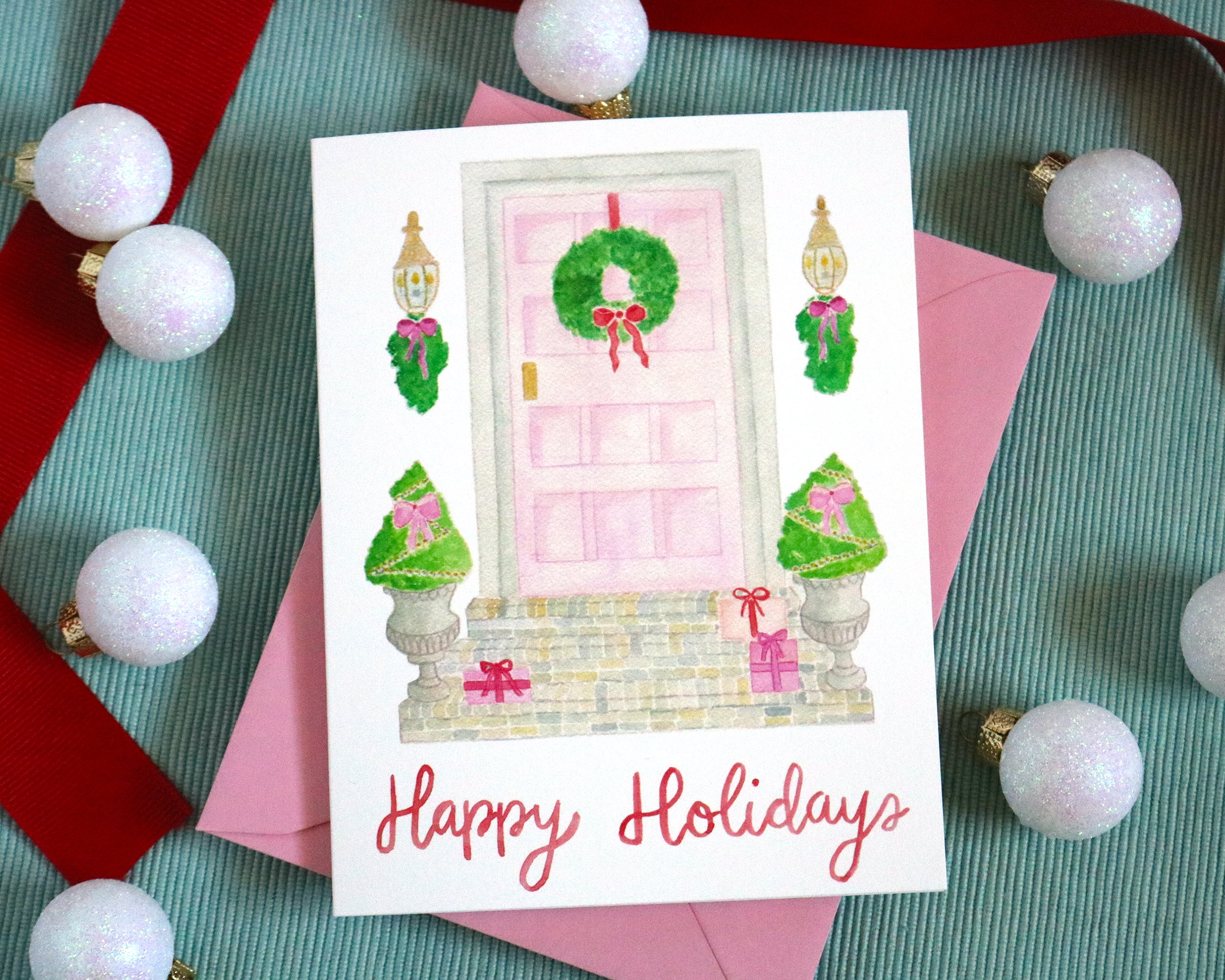 Our greeting card features our watercolor painting of a festive pink front door with the words “Merry Christmas” in our signature calligraphy. Displayed with red ribbon and white ornaments.