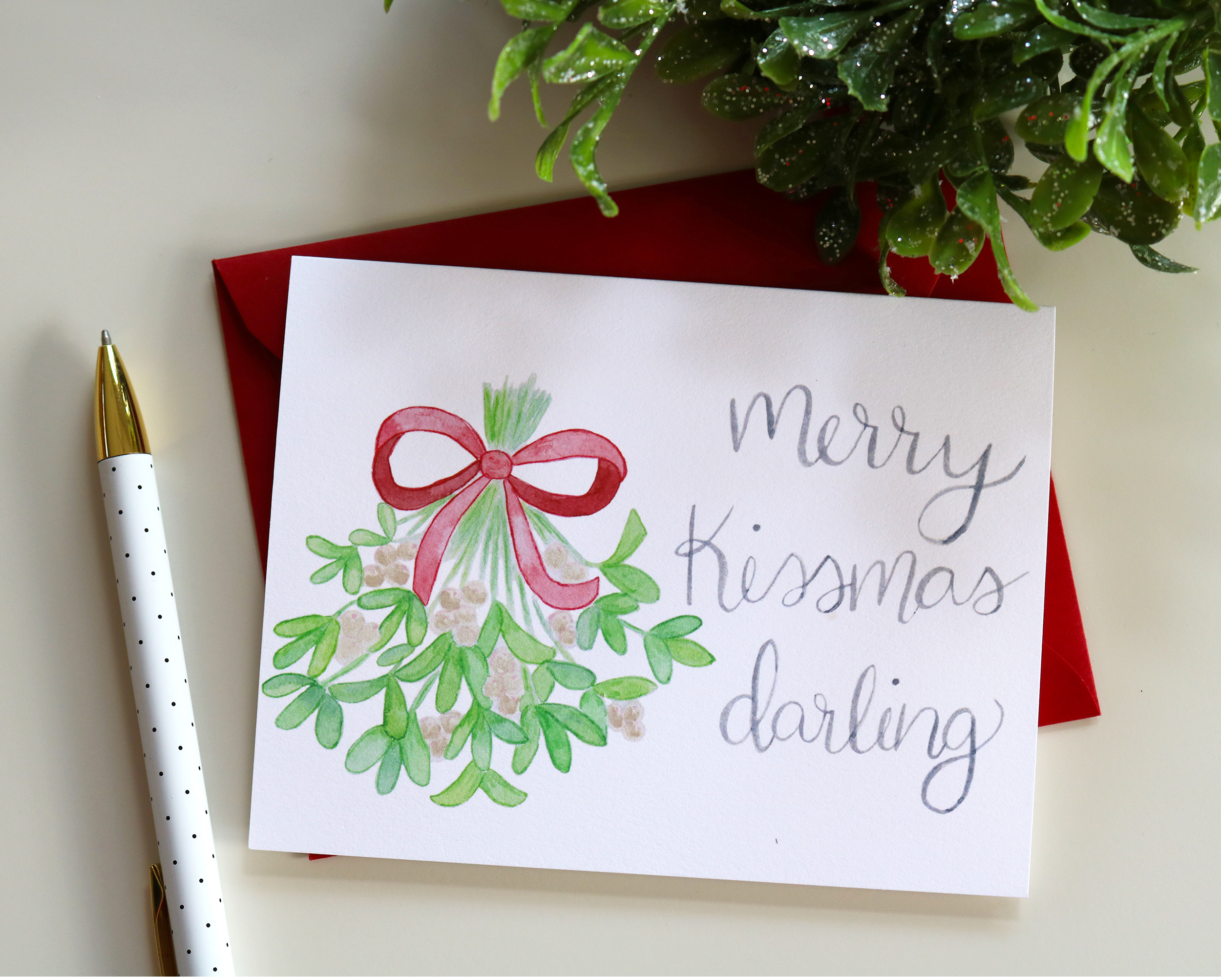 Our card features our watercolor painting of mistletoe tied with a red ribbon and iridescent berries next to the words “Merry Kissmas Darling” in our signature calligraphy. Comes with a red envelope.