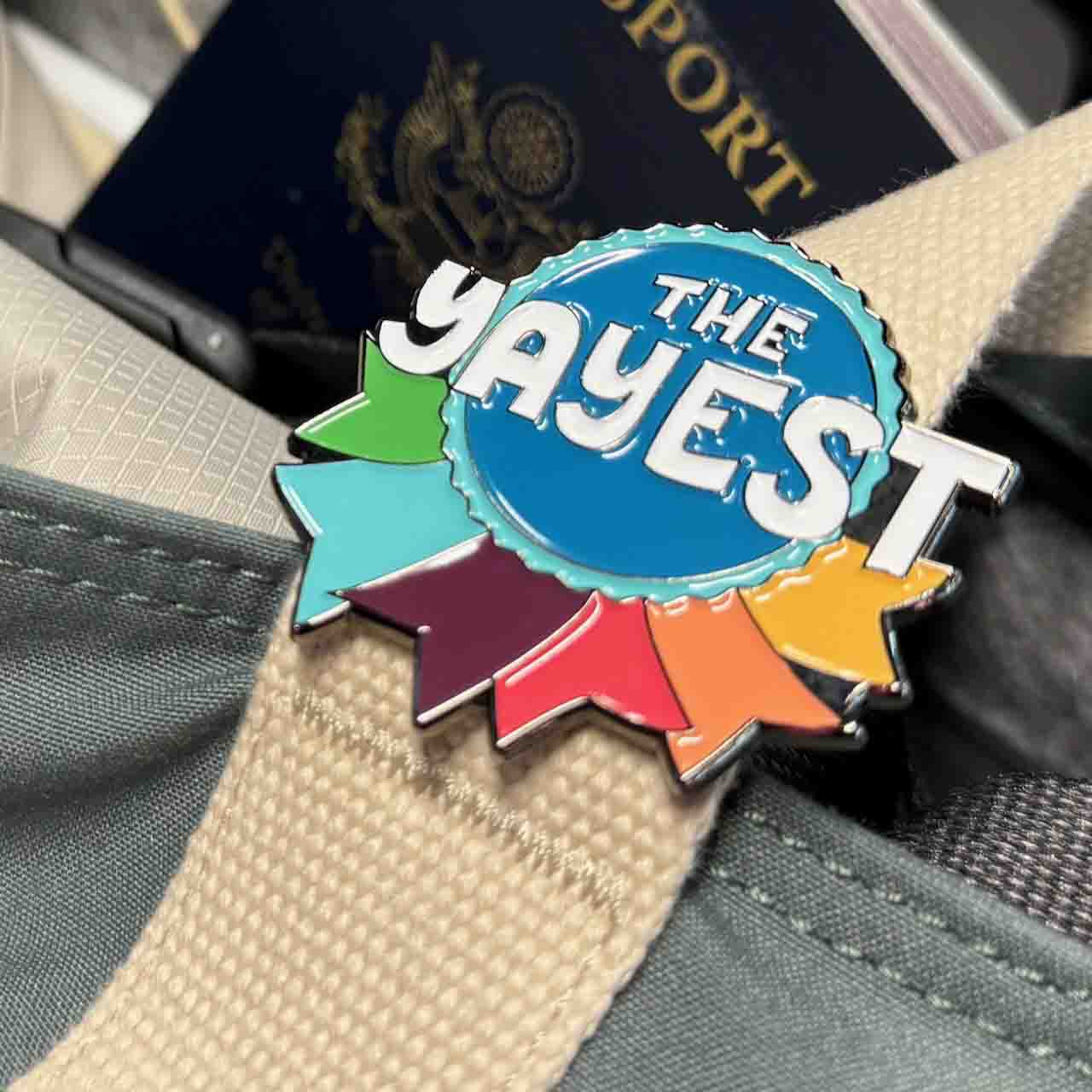 An enamel pin with a rainbow ribbon design that says "The Yayest"
