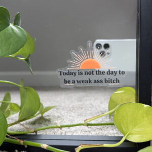 Window cling affixed to a mirror with an orange sun and black text that says today is not the day to be a weak ass bitch