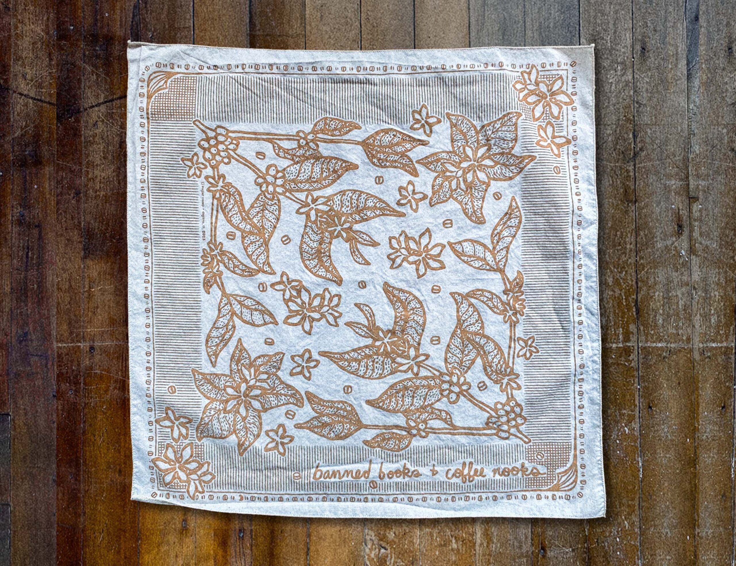 Sugar Camp Banned Books & Coffee Nooks Bandana in cream, lying flat on distressed wooden floor. Motif of coffee plants, coffee beans, and worn book edges.