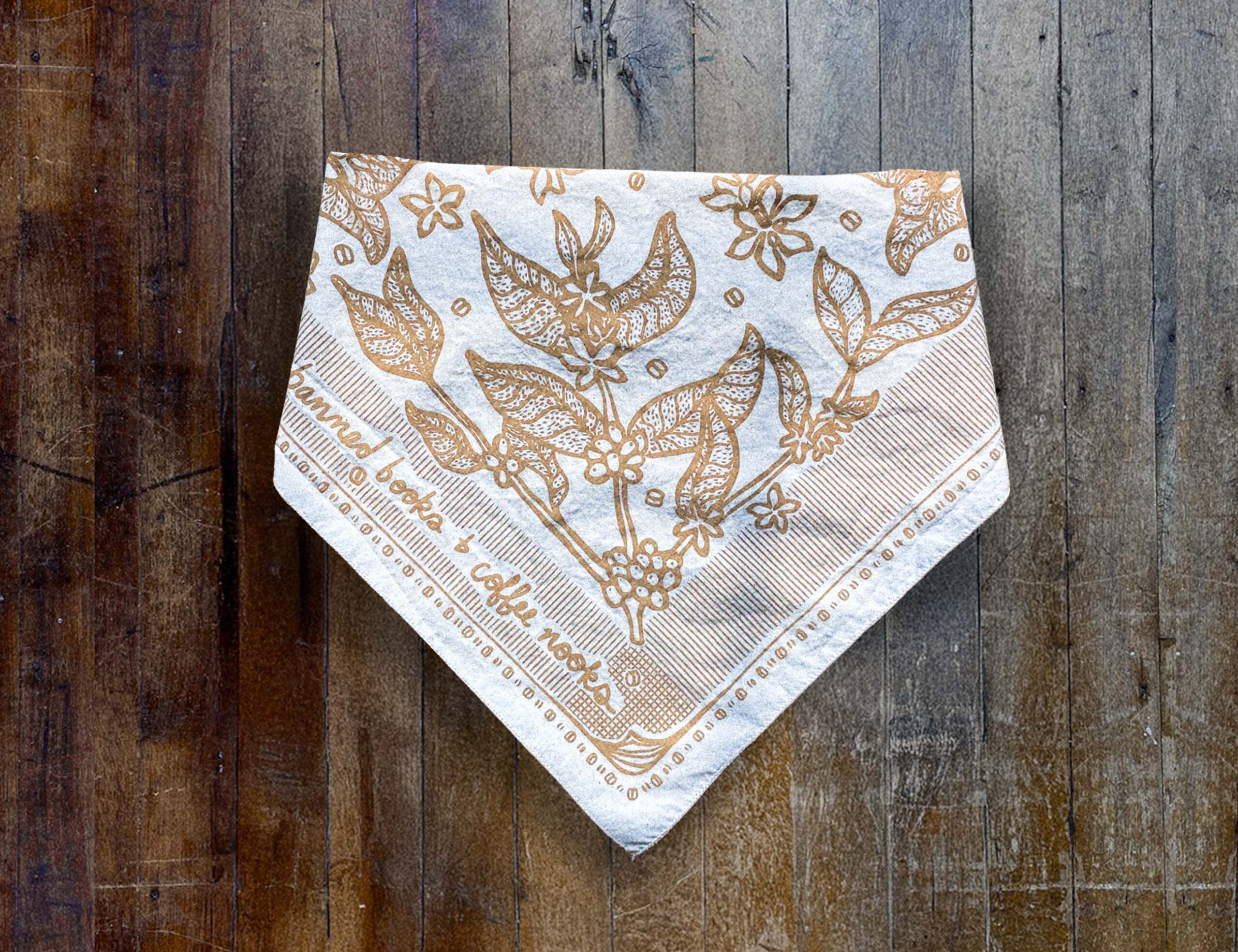 Sugar Camp Banned Books & Coffee Nooks Bandana in cream, lying folded on distressed wooden floor. Motif of coffee plants, coffee beans, and worn book edges.