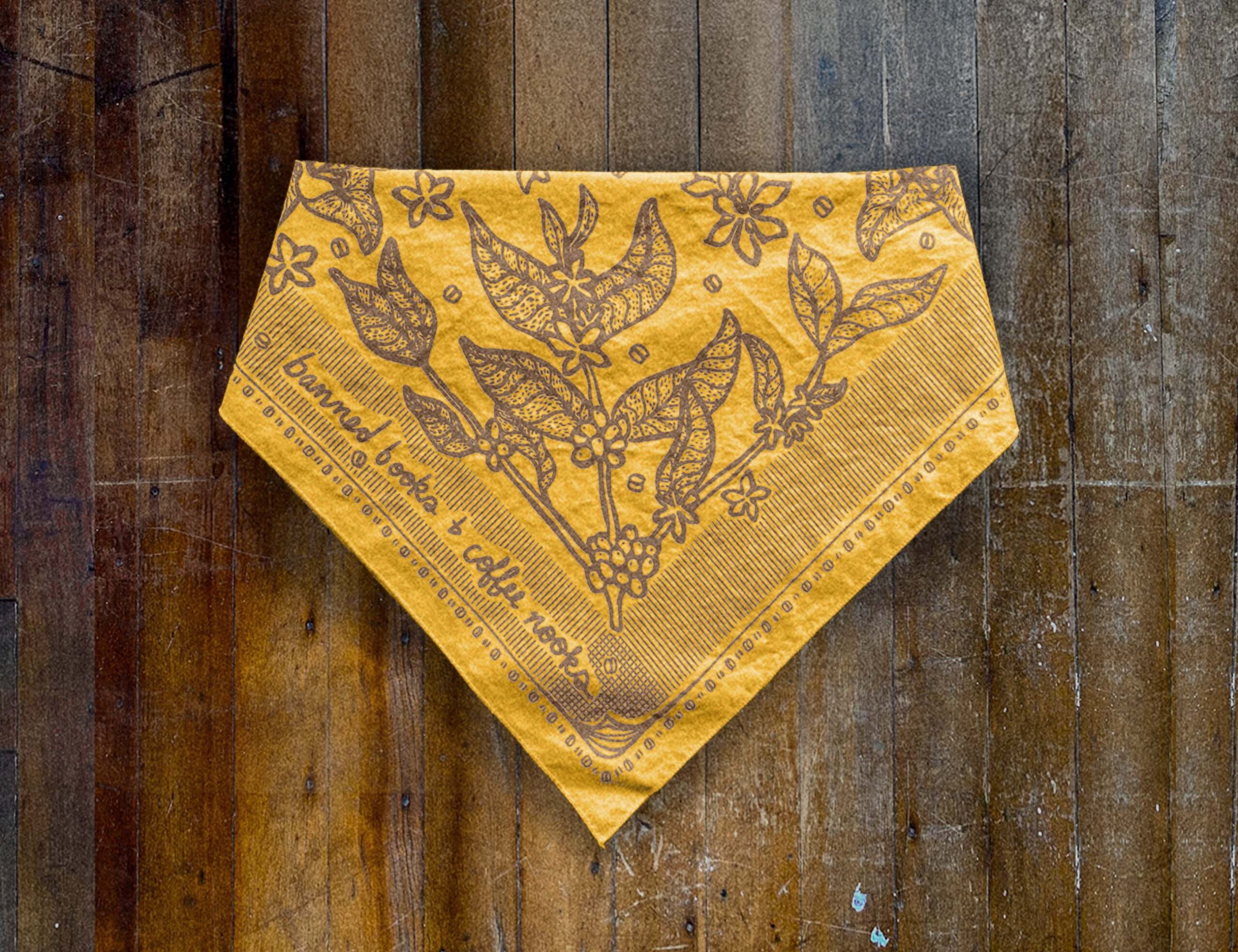 Sugar Camp Banned Books & Coffee Nooks Bandana in groovy gold, lying folded on distressed wooden floor. Motif of coffee plants, coffee beans, and worn book edges.