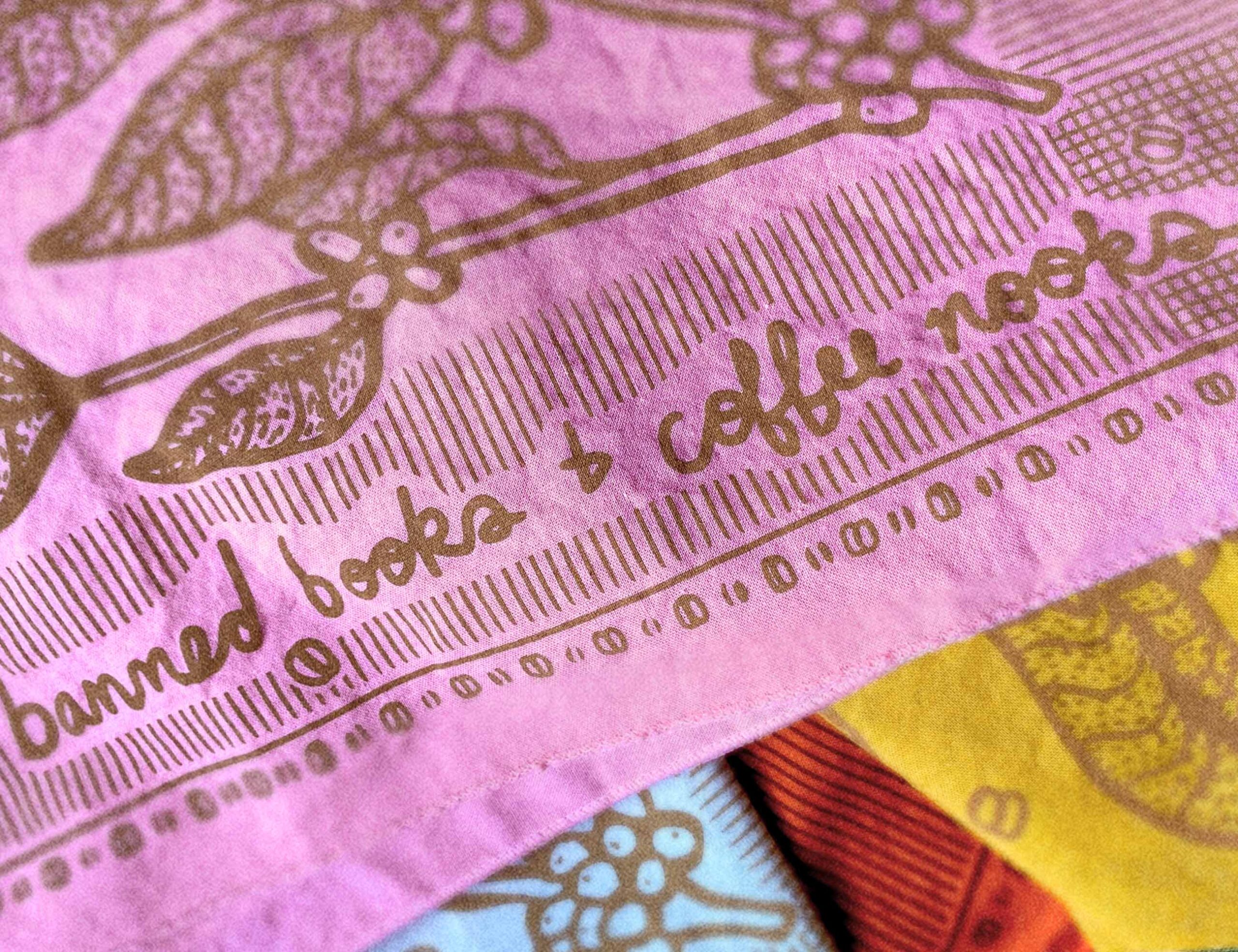 sugar camp banned books and coffee nooks bandana in flower power pink in a pile over other bandanas. has a motif of coffee plants and worn book edges.