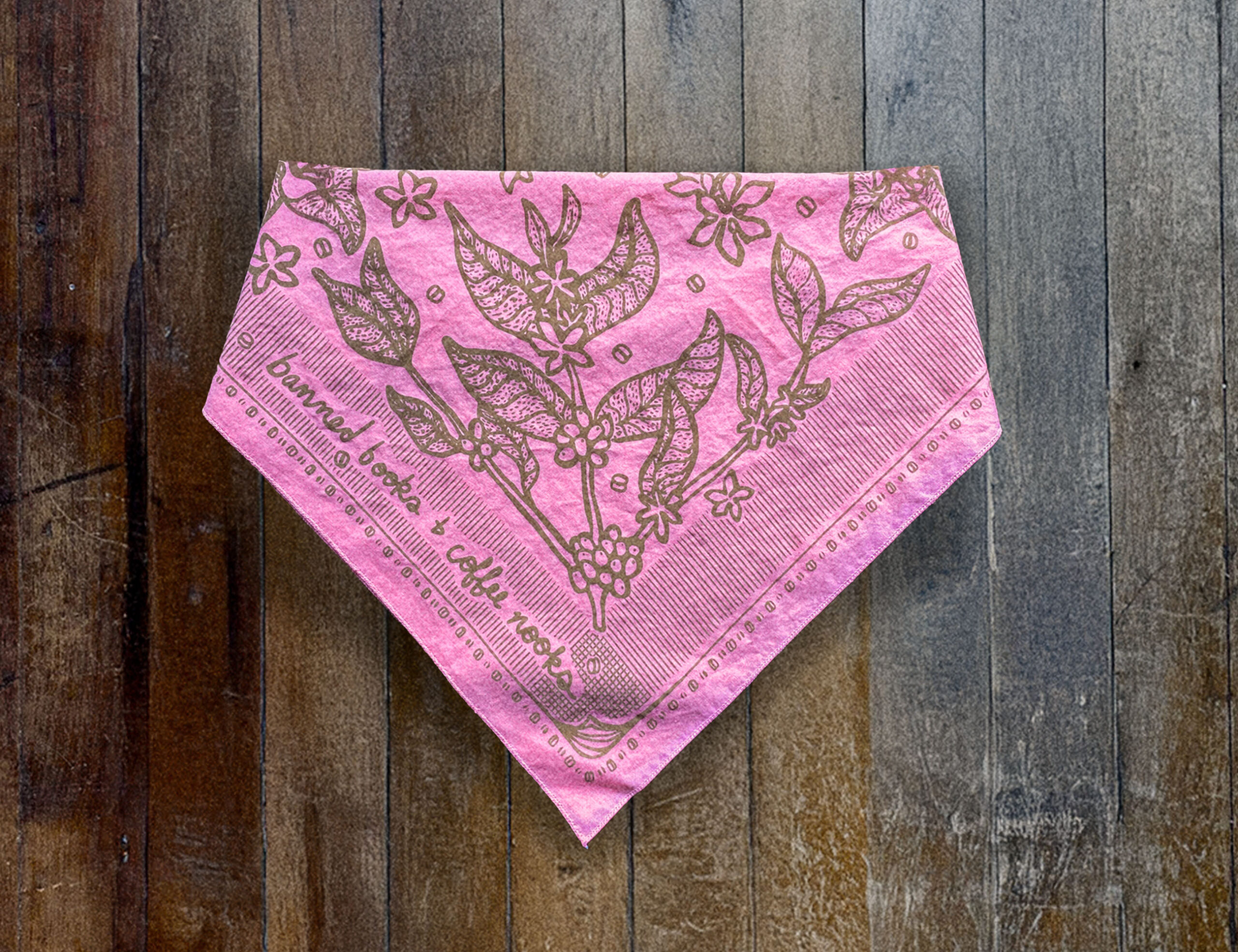 Sugar Camp Banned Books & Coffee Nooks Bandana in pink, lying folded on distressed wooden floor. Motif of coffee plants, coffee beans, and worn book edges.