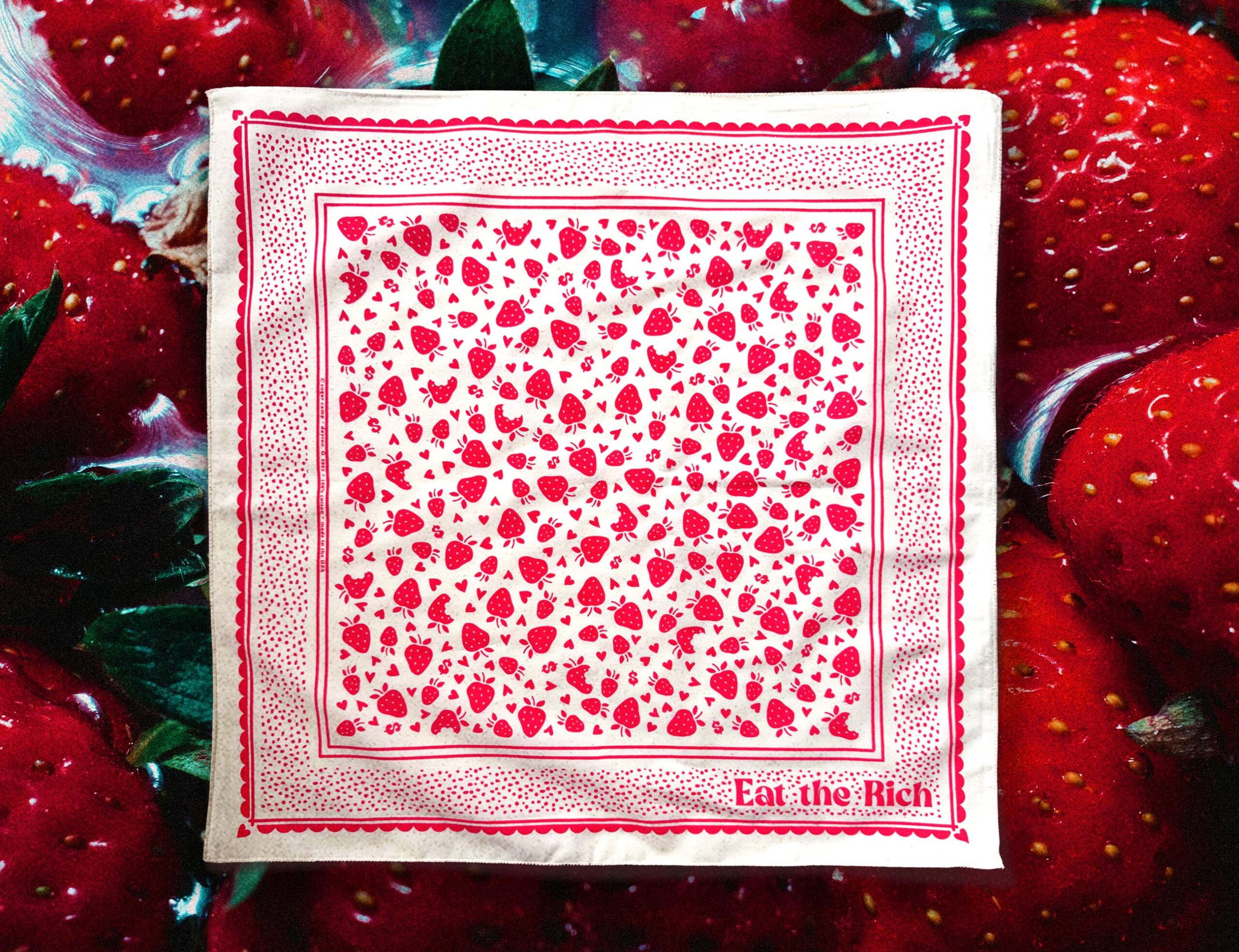 Cream “Eat the Rich” bandana with red strawberry, heart, and dollar sign print over a photo of red strawberries in water