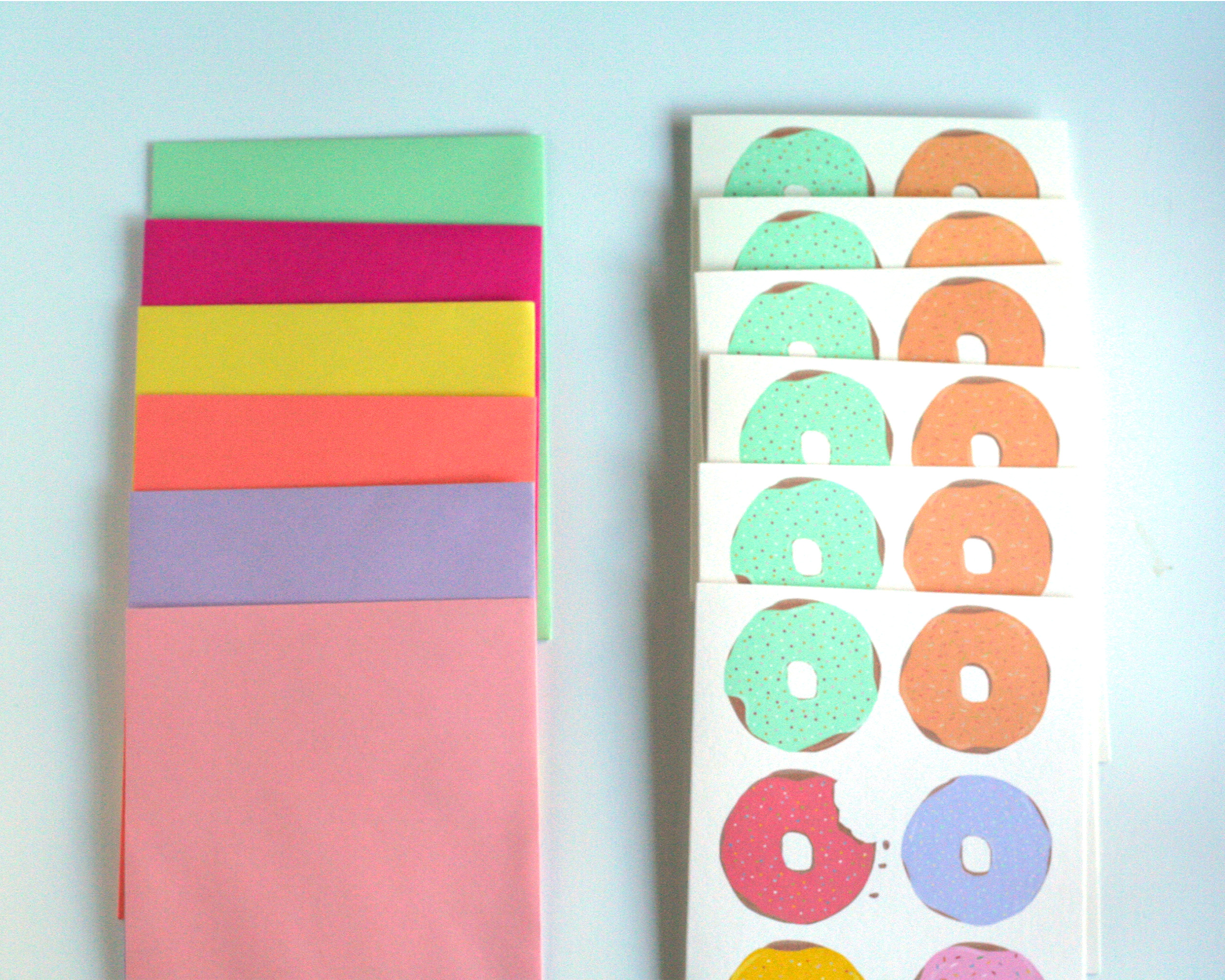 Greeting Card with 6 illustrated donuts with frosting in pastel rainbow colors on white background. Pictured next to a rainbow of envelopes.