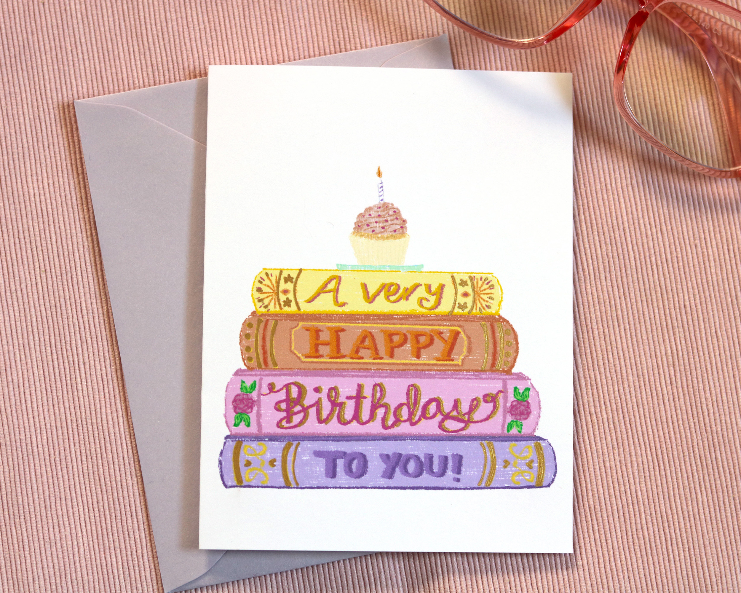 Greeting card with illustrated stack of books with words "wishing you a happy birthday" and a cupcake on top.