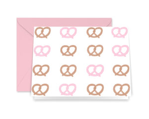 Photo of our Illustrated Pink Pretzel Greeting Card and pink envelope. The card features a pattern of illustrated pink and brown pretzels on a white background.