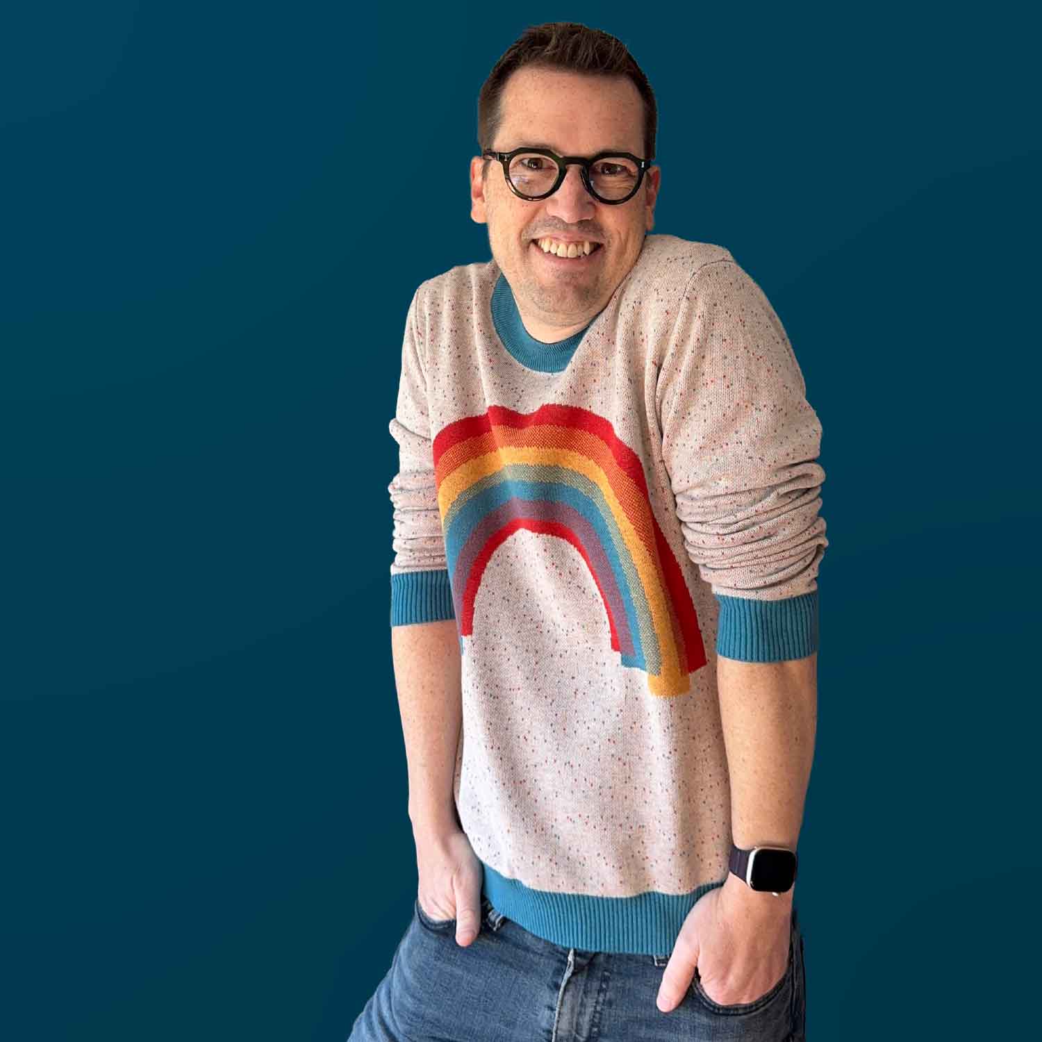 A portrait of Jedd wearing a white sweater with a rainbow design on the front