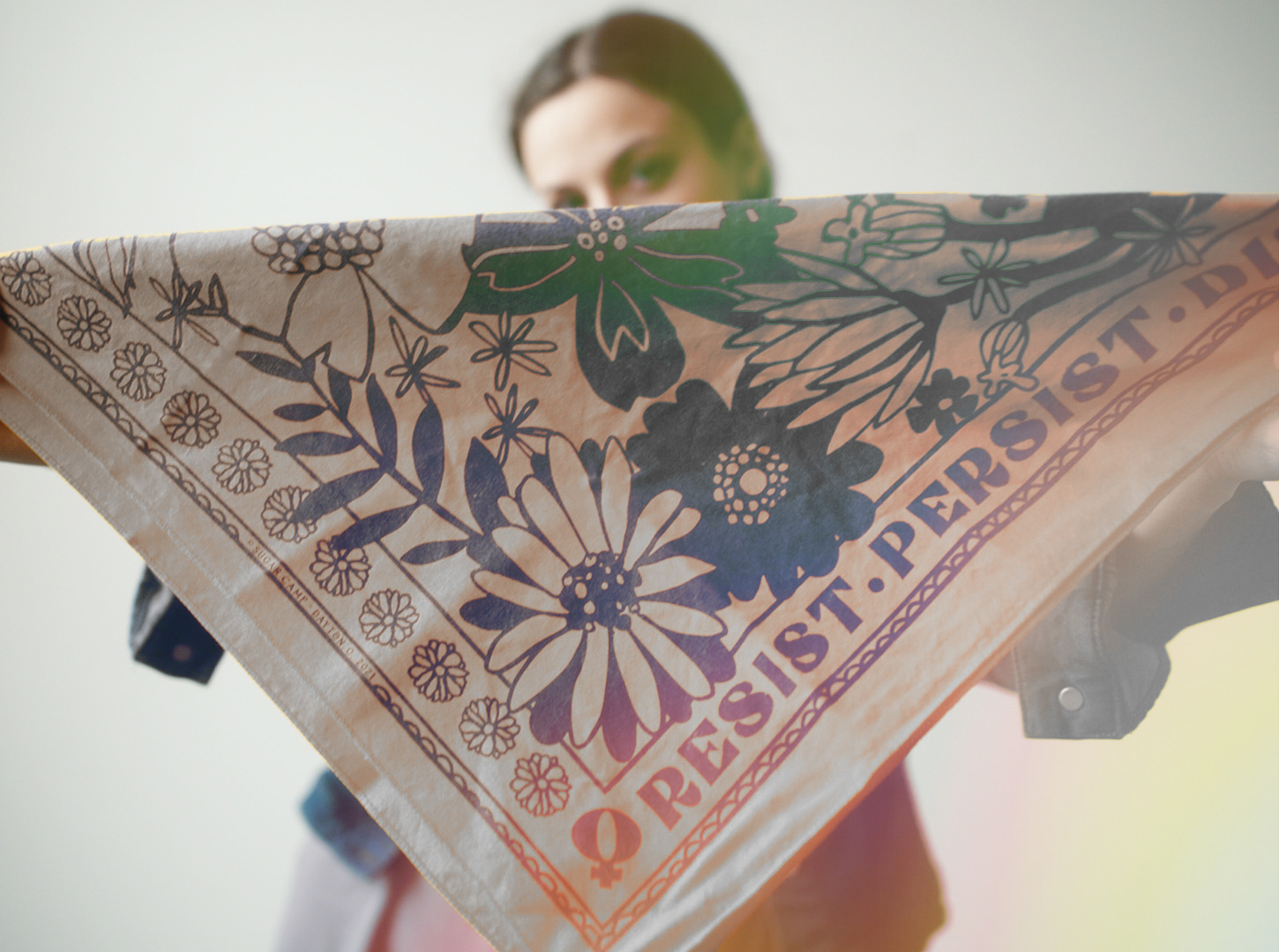 young woman holds up sugar camp Resist Persist Dissent bandana in tan sand.