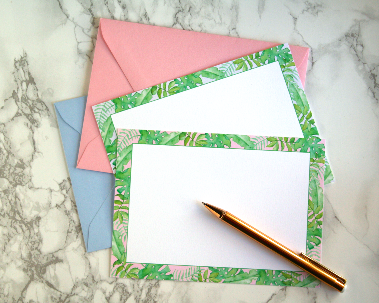 A photo of our Watercolor Tropical Leaves Flat Notecard Stationery Set in both pastel pink and blue with coordinating envelopes on a marble background next to a gold pen.