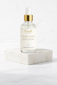 Luxury room and linen spray bottle featuring Camellia Lux Scents Exotic Lemon fragrance in 4 oz clear glass bottle with ivory and gold label, gold metallic spray nozzle, photographed on white marble platform against neutral background