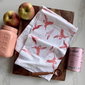 White cotton tea towel with a pink hummingbird print, styled on a wooden cutting board & kitchen counter with apples & pink containers