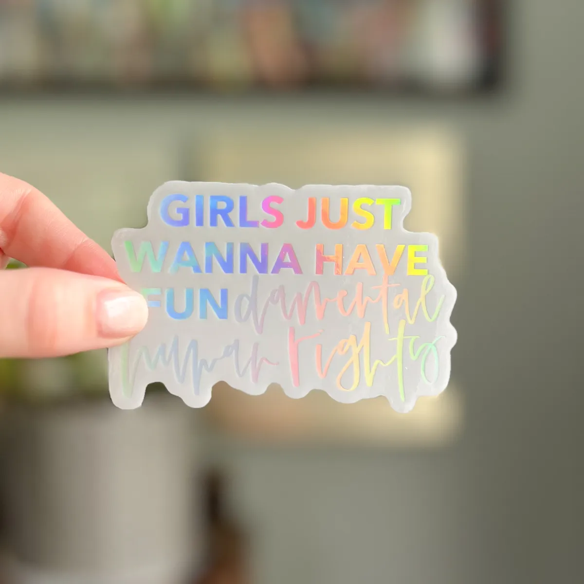 a white sticker with the quote "girls just wanna have fundamental human rights" in holographic block letters and modern calligraphy
