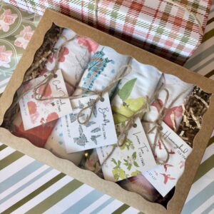 Kraft brown box with a clear window containing four white towels with various colorful prints, rolled and tied with twine and a square hang tag