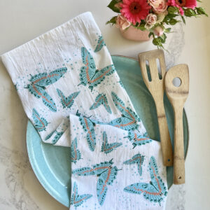 Blue butterfly print tea towel, styled on a blue plate and kitchen counter with wooden cooking utensils and pink flowers